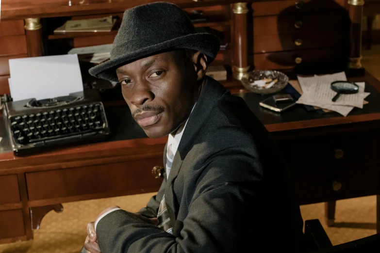 a man sitting at a desk with a typewriter, an album cover, inspired by George Bain, harlem renaissance, lance reddick, a suited man in a hat, close up portrait photo, taken in the late 2000s