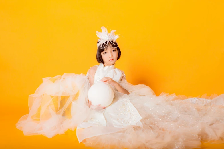 a little girl in a white dress holding a white ball, an album cover, by Ayami Kojima, pexels contest winner, yellow backdrop, elegant glamourous cosplay, headpiece headpiece headpiece, 15081959 21121991 01012000 4k