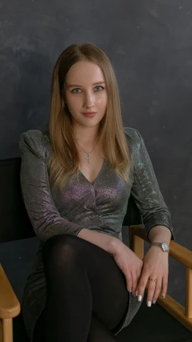 a woman sitting in a chair in front of a black wall, wearing a designer top, pokimane, grey and silver, low quality photo