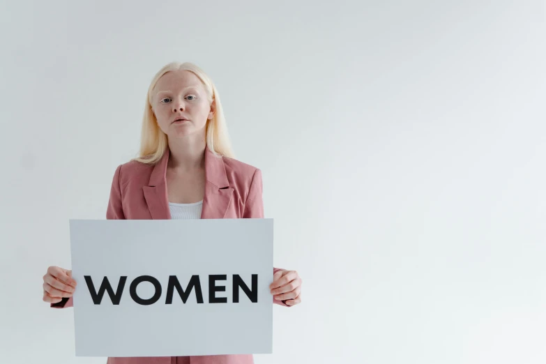 a woman holding a sign that says women, by Emma Andijewska, trending on pexels, on a pale background, human staring blankly ahead, with a white complexion, female gigachad