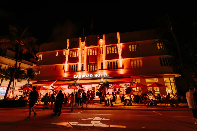 a group of people walking in front of a hotel at night, orange and red lighting, art deco buildings, festival, lizzo