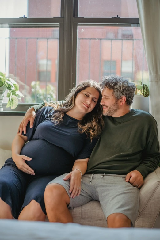 a man and woman sitting on a couch in front of a window, pexels contest winner, renaissance, maternity feeling, grey, official screenshot, brett goldstein