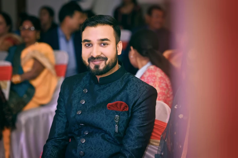 a man sitting in front of a crowd of people, inspired by Saurabh Jethani, pexels contest winner, hurufiyya, with an elegant smile, formal attire, headshot profile picture, with a small beard