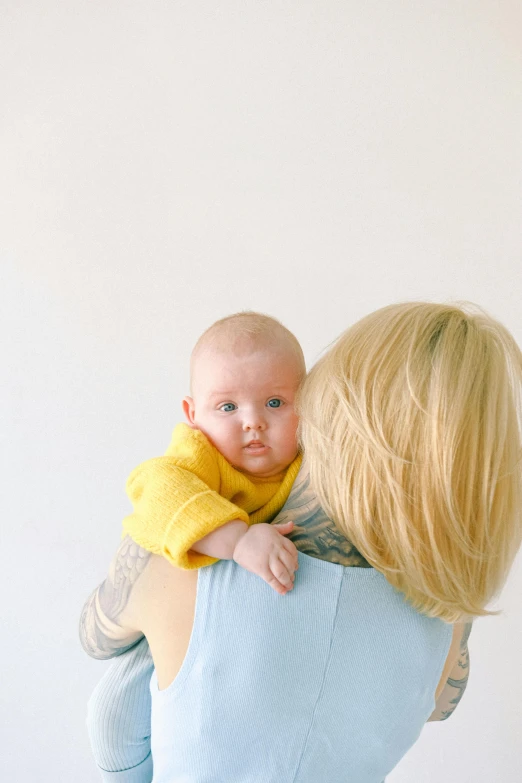 a woman holding a baby in her arms, by Ellen Gallagher, pexels, yellow clothes, looking from shoulder, on a pale background, blond