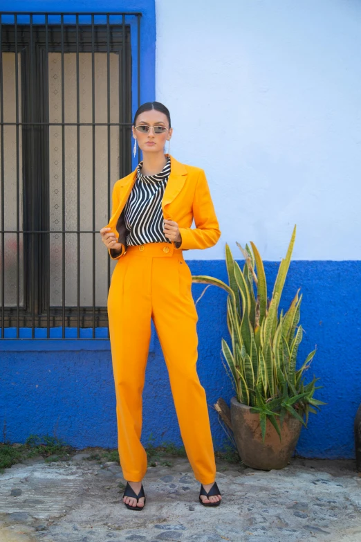 a woman standing in front of a blue building, in a gold suit, wearing a pinstripe suit, colors: yellow, lorena avarez