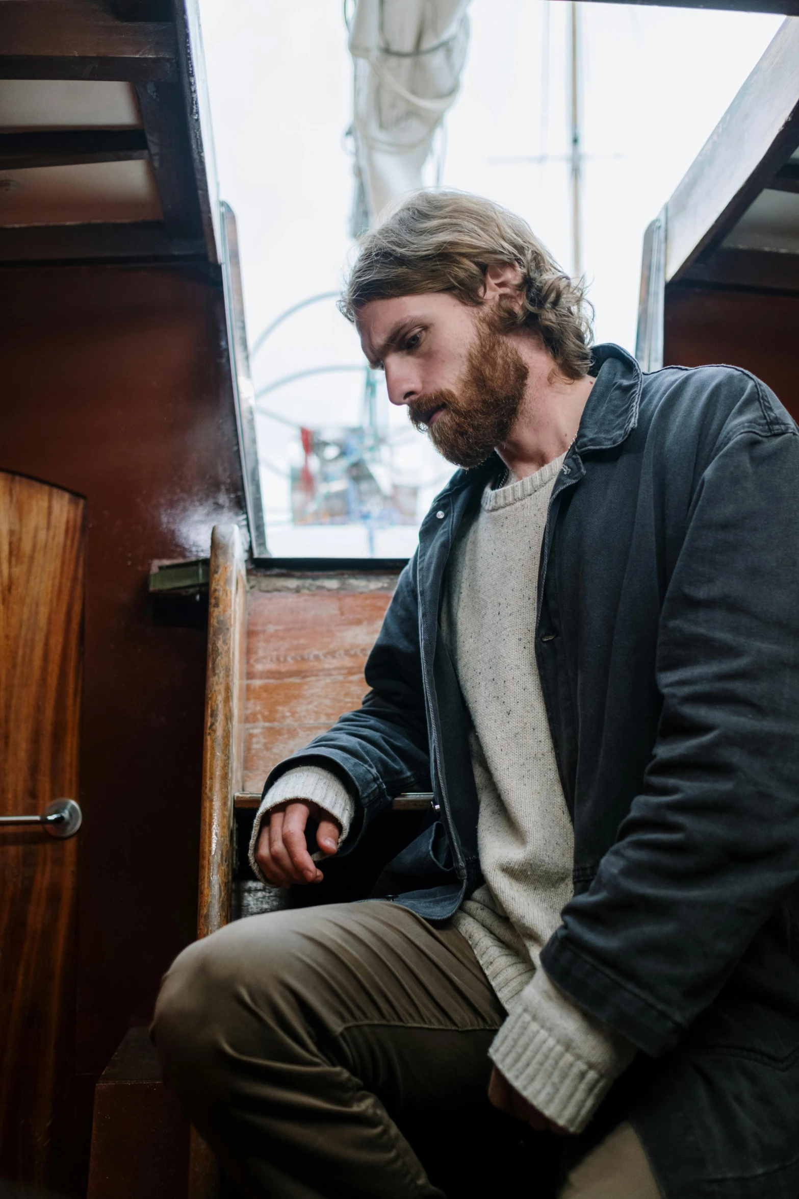 a man sitting on top of a wooden bench, wearing a cardigan, on a boat, caramel. rugged, smoking a bowl of hash together