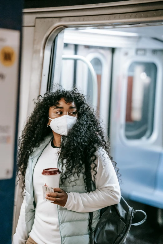 a woman wearing a face mask while holding a cup of coffee, trending on pexels, afrofuturism, inside of a metro train, sza, polluted, curly haired