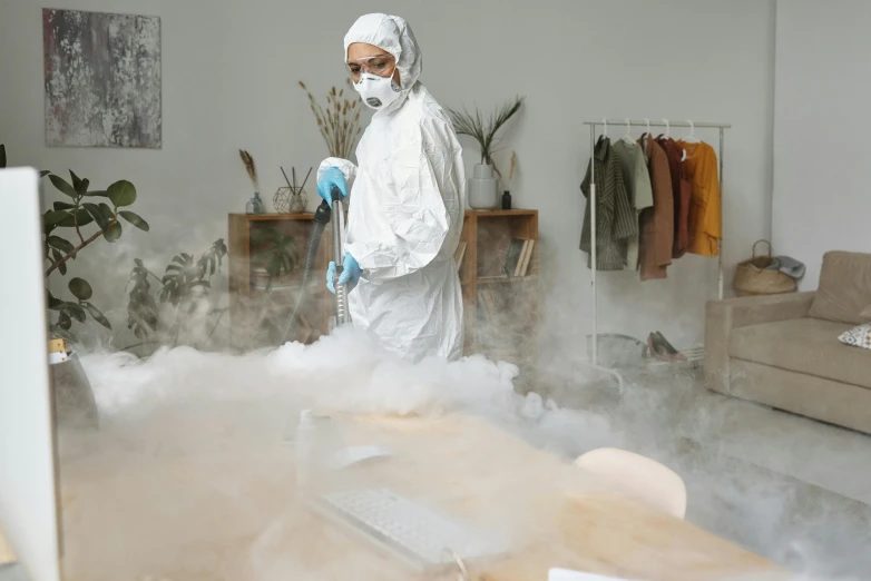 a man in a white hazmat suit standing in a living room, by Julia Pishtar, pexels contest winner, dry ice, working in her science lab, misting, wearing a plug suit