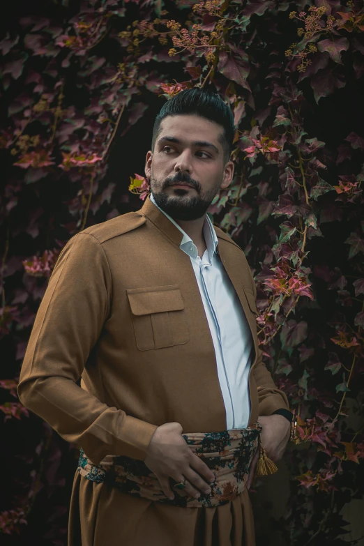 a man standing in front of a wall with flowers, an album cover, by Alejandro Obregón, pexels contest winner, wearing elegant casual clothes, caramel. rugged, assyrian, portait photo profile picture