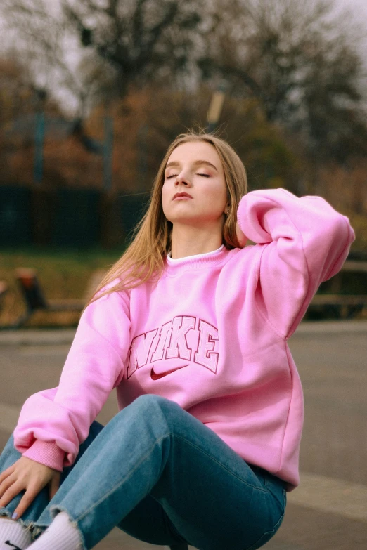 a woman sitting on a skateboard in a parking lot, an album cover, inspired by Elsa Bleda, trending on pexels, hyperrealism, wearing a pastel pink hoodie, large nike logo, sitting on a park bench, frown fashion model