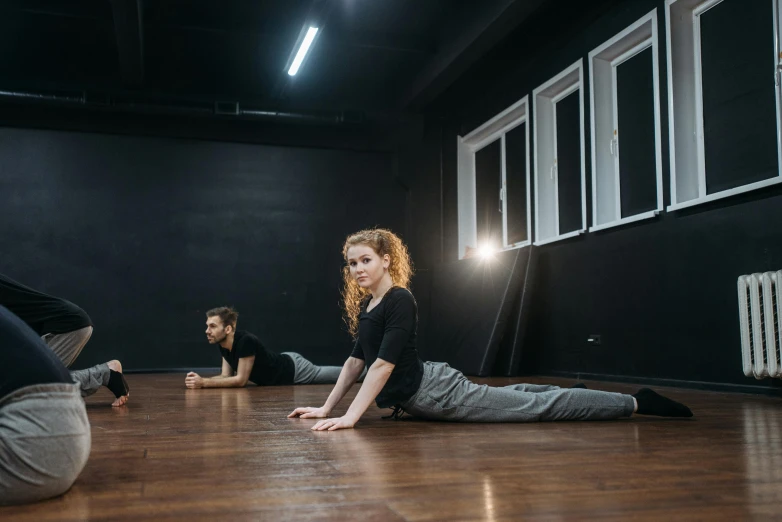 a group of people sitting on the floor in a room, doing splits and stretching, in an empty black room, profile image, square