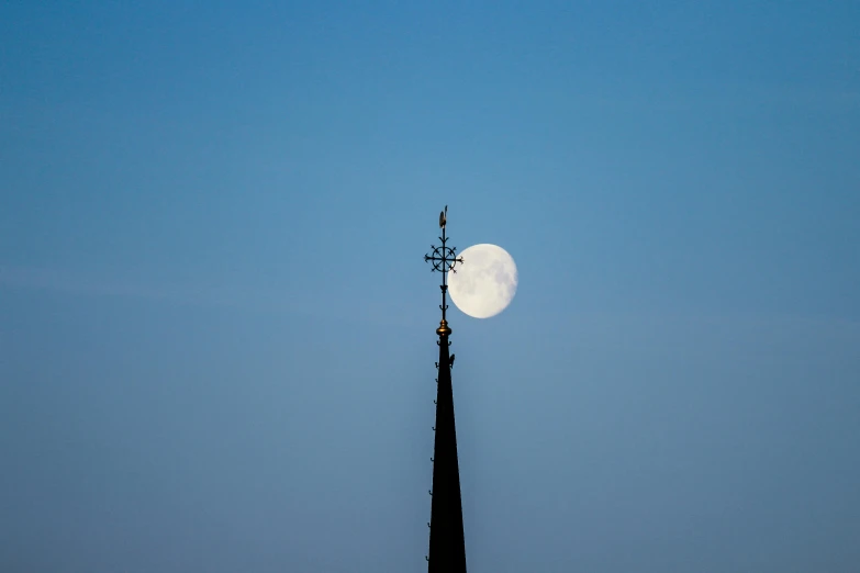 a cross on top of a steeple with a full moon in the background, by Peter Churcher, unsplash, minimalism, shot on sony a 7 iii, light blue clear sky, spire, evening sunlight