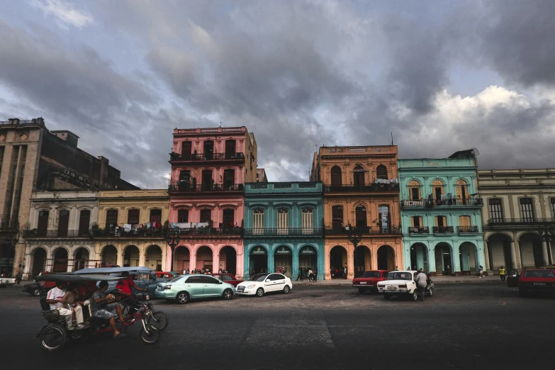 a group of cars driving down a street next to tall buildings, a colorized photo, pexels contest winner, hyperrealism, cuban setting, ancient buildings, square, colorful with red hues