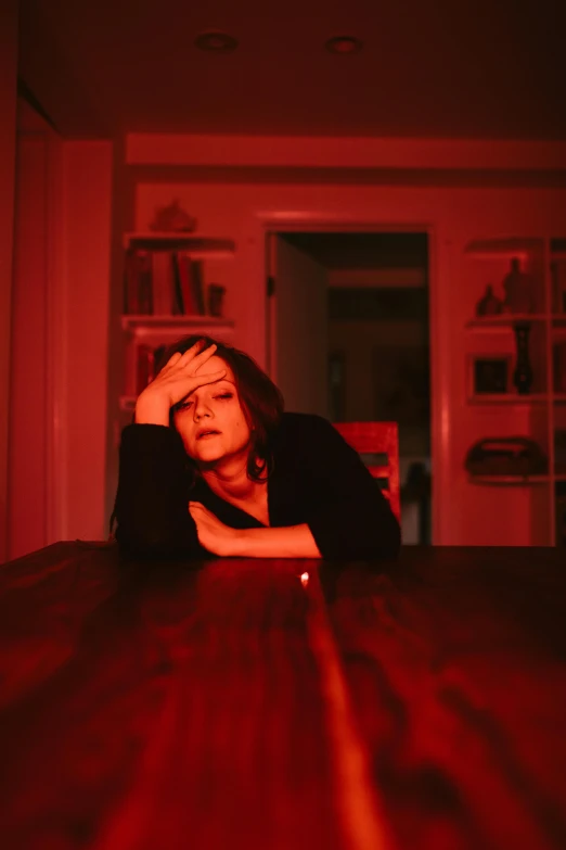 a woman sitting at a table with her head in her hands, an album cover, inspired by Nan Goldin, pexels, blood aura red light, clarice starling, unhappy, standing in a dimly lit room