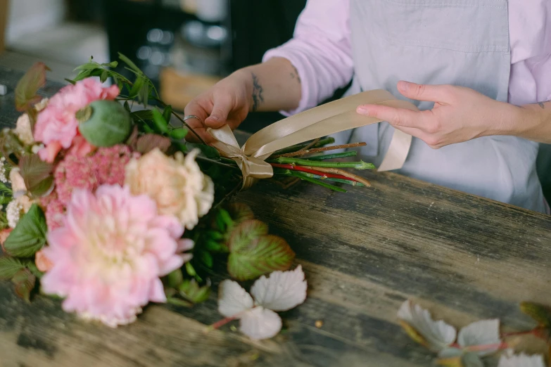 a woman holding a bunch of flowers on top of a wooden table, insisted on cutting in line, with intricate details, brown and pink color scheme, bending down slightly