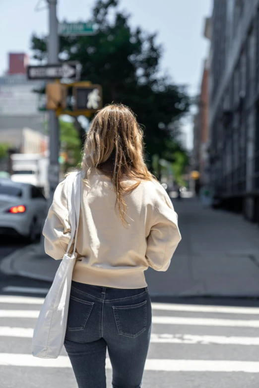 a woman walking across a crosswalk in a city, by Nina Hamnett, happening, beige hoodie, showing her shoulder from back, watching new york, on a sunny day