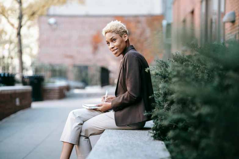 a woman sitting on a ledge reading a book, a portrait, by Alice Mason, pexels contest winner, short blonde afro, business attire, portrait androgynous girl, brown jacket with long sleeves