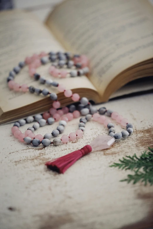 an open book sitting on top of a wooden table, gemstone necklace, pink and grey muted colors, figure meditating close shot, beads of sweat