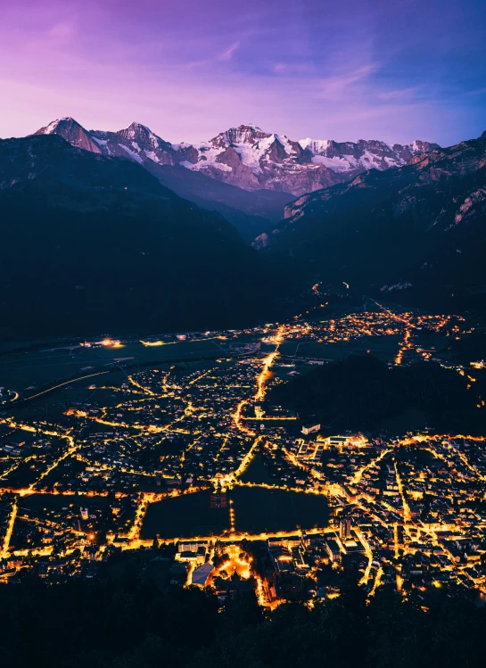 a city lit up at night with mountains in the background, in the swiss alps, top - down photograph, 4k image”, grain”