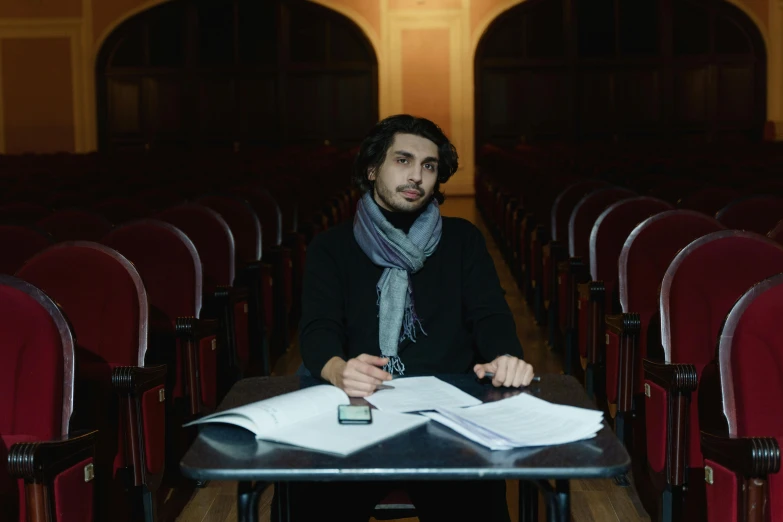 a man sitting at a table with papers in front of him, an album cover, inspired by Ismail Acar, pexels contest winner, academic art, standing in a large empty hall, man wearing a closed cowl, graduation photo, looking straight to camera