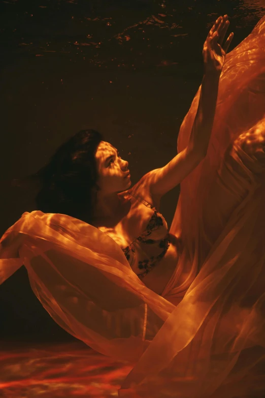 a woman floating in a body of water, orange glow, showstudio, draped in gold, in a underwater horror scene