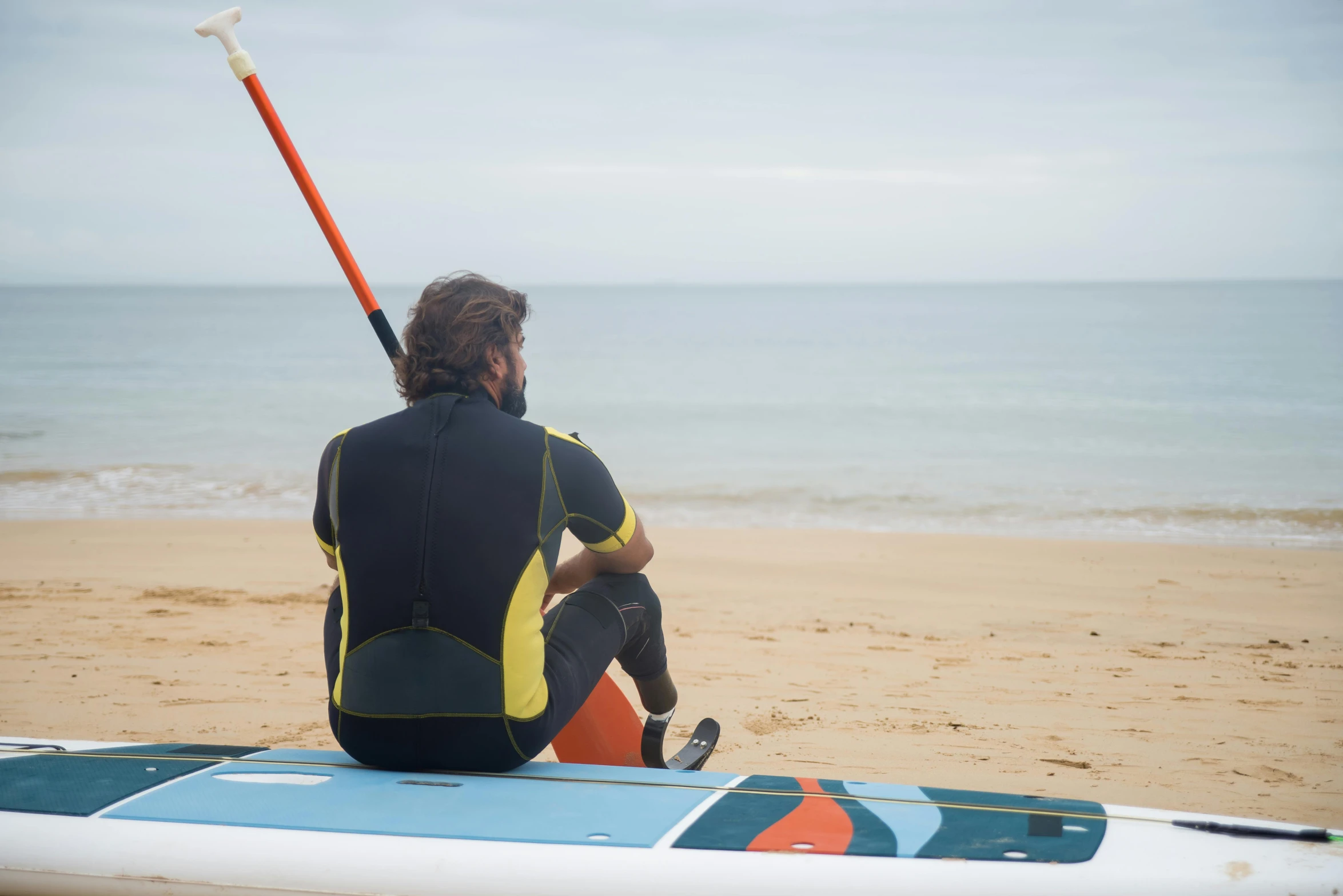 a man sitting on top of a surfboard on a beach, gazing at the water, neoprene, heavy lines, with a sleek spoiler