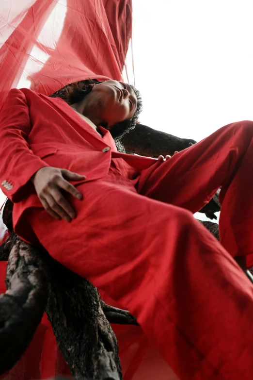 a woman sitting on top of a bed covered in a red sheet, by Matija Jama, wearing a worn out suit, sitting in a tree, demobaza, dark skinned