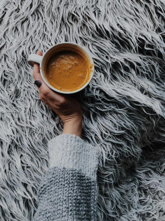 a person holding a cup of coffee on a furry rug, by Lucia Peka, high quality image
