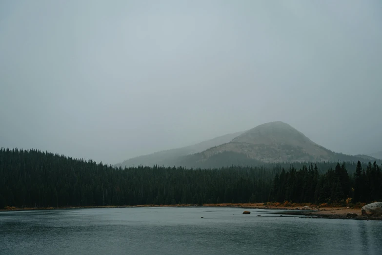 a body of water with a mountain in the background, under a gray foggy sky, boreal forest, low quality photo