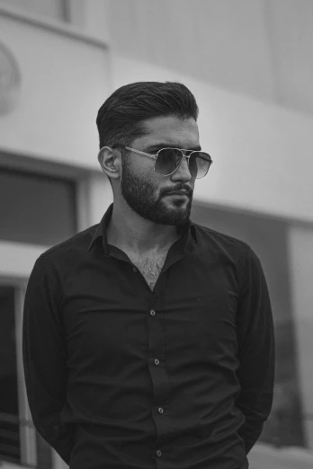 a man with a beard standing in front of a building, a black and white photo, by Ismail Acar, pexels contest winner, a handsome man，black short hair, with sunglass, portait photo profile picture, handsome young man