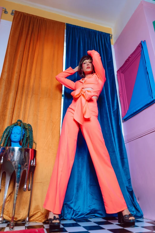 a woman standing on a black and white checkered floor, an album cover, inspired by David LaChapelle, trending on pexels, maximalism, wearing an orange jumpsuit, leaning on door, holographic suit, flowing salmon-colored silk