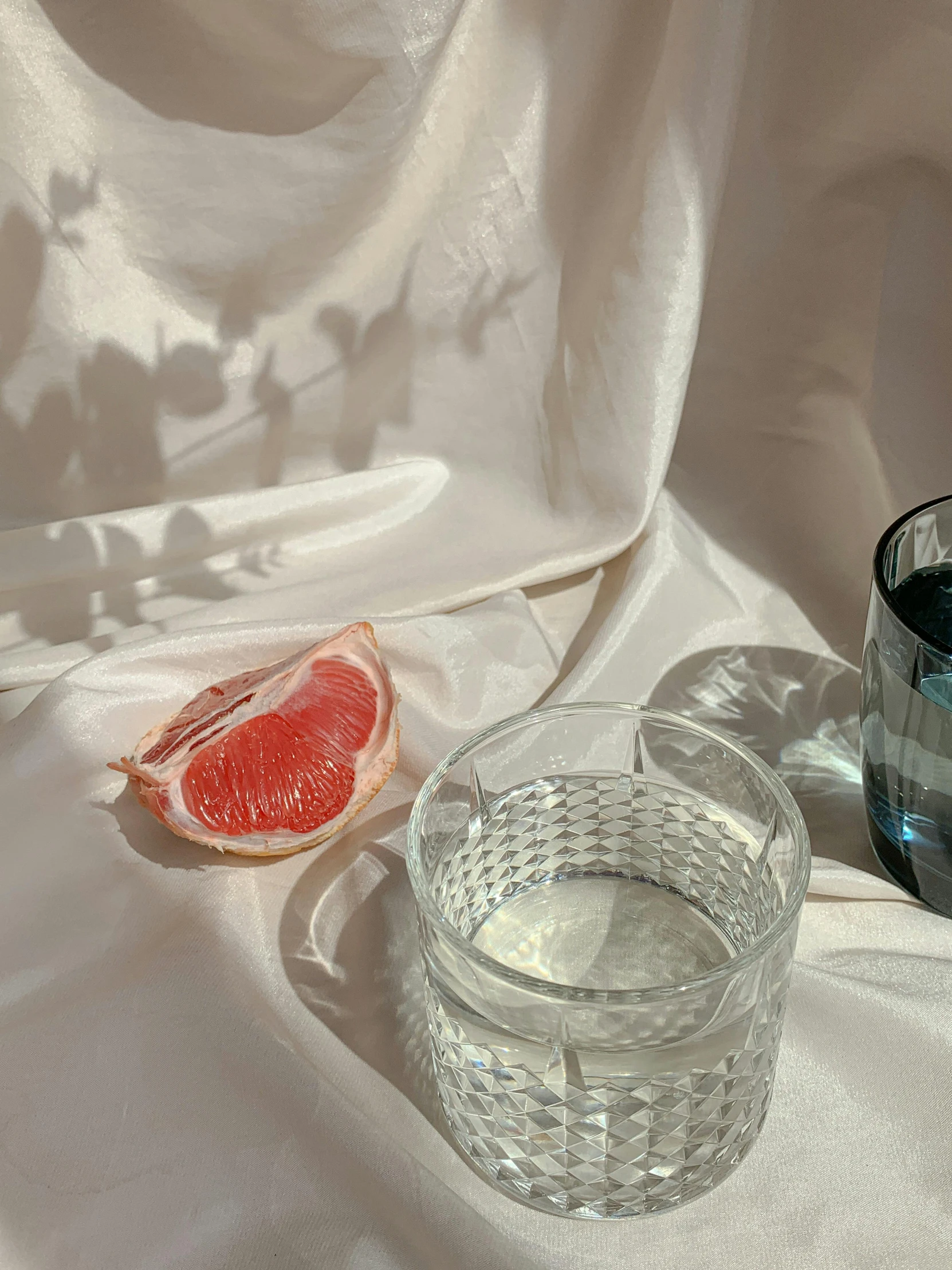 a glass of water sitting next to a grapefruit, by Nathalie Rattner, white and pink cloth, low quality photograph, ilustration, translucent glass shine