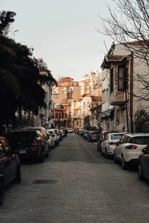 a street lined with parked cars next to tall buildings, by Ismail Acar, ancient city streets behind her, high quality image