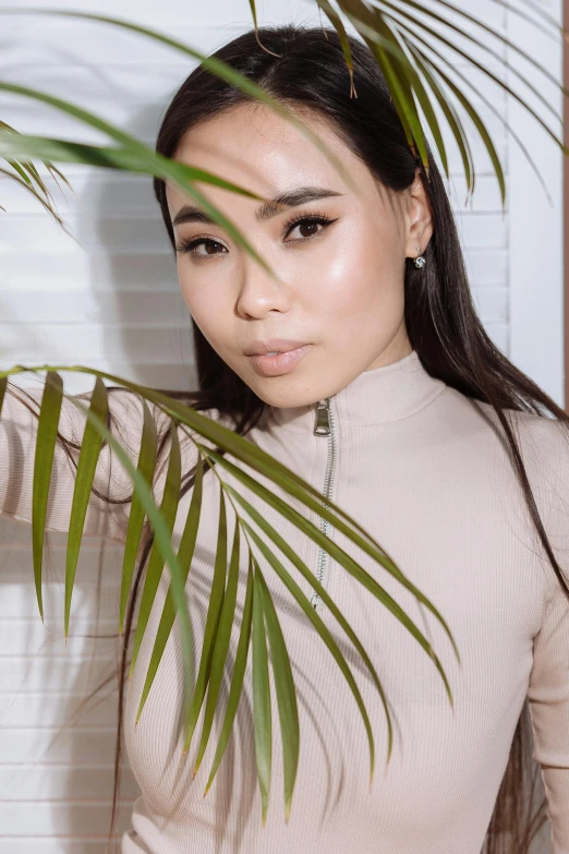 a woman standing next to a palm tree, an album cover, inspired by Ruth Jên, trending on pexels, young cute wan asian face, wearing turtleneck, beige, a high angle shot