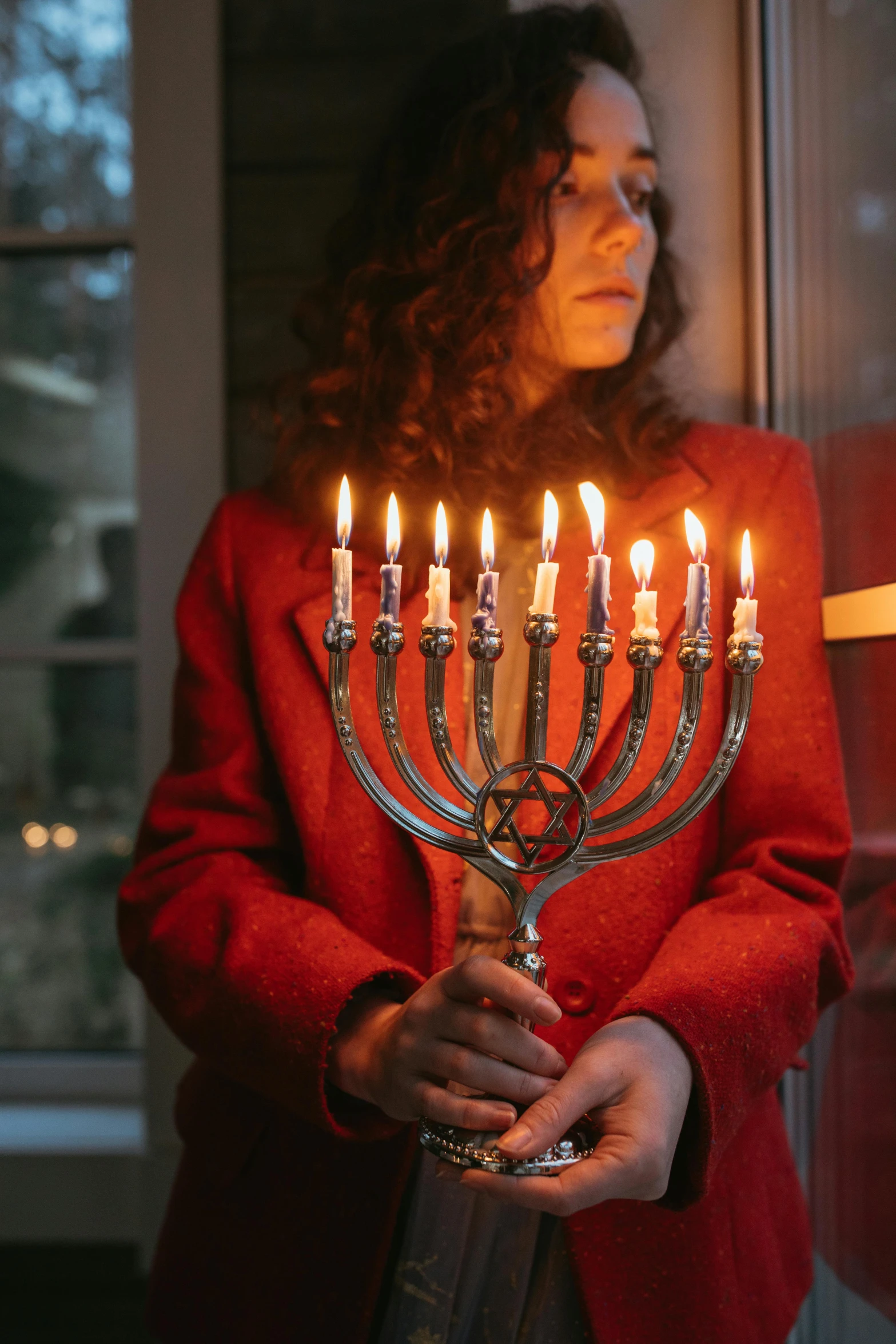 a woman holding a menorah in front of a window, pexels, (1 as december, wearing red jacket, waves of lights, jen atkin