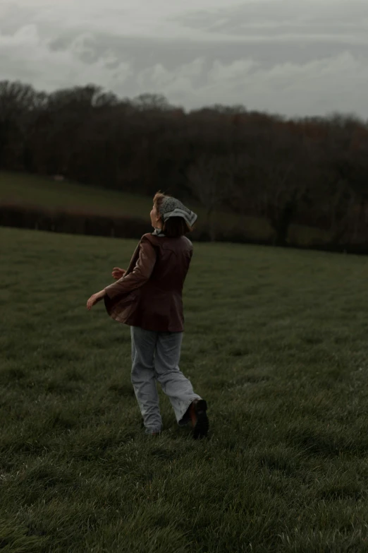 a person in a field flying a kite, an album cover, unsplash, wearing a brown leather coat, walking at the garden, lena oxton, gloomy mood