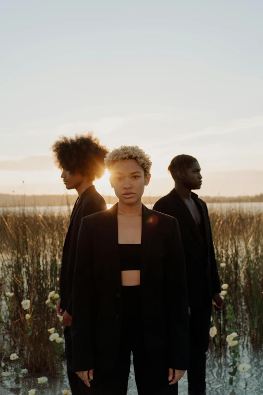 a group of people standing next to each other in a field, an album cover, by Winona Nelson, unsplash, afrofuturism, short blonde afro, near a lake, trio, woman in black robes