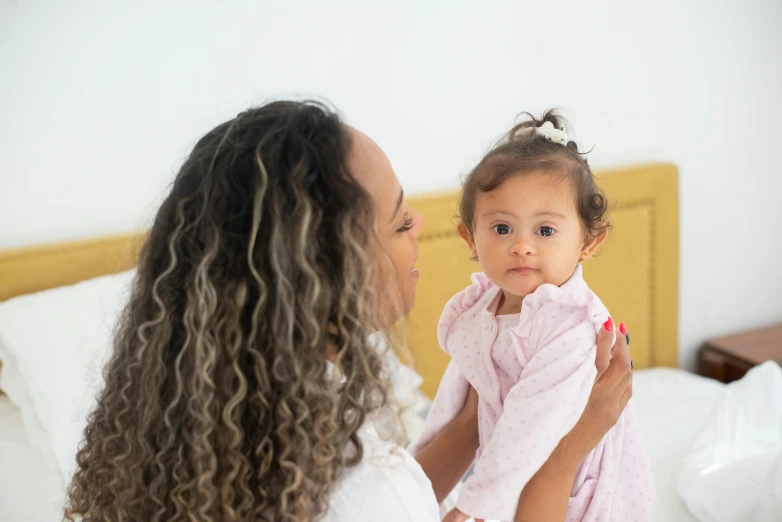 a woman holding a baby on top of a bed, pexels contest winner, hurufiyya, mixed-race woman, looking partly to the left, wearing pajamas, curls on top