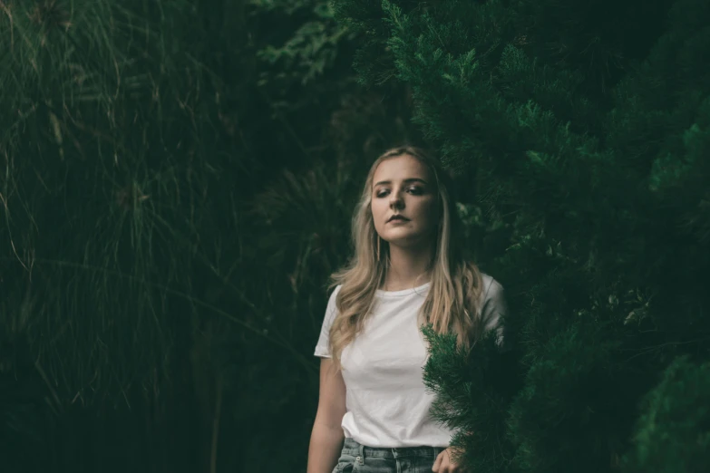 a woman standing in front of a tree, inspired by Elsa Bleda, pexels contest winner, realism, portrait of kim petras, dark green tones, dressed in a white t-shirt, unsplash photography