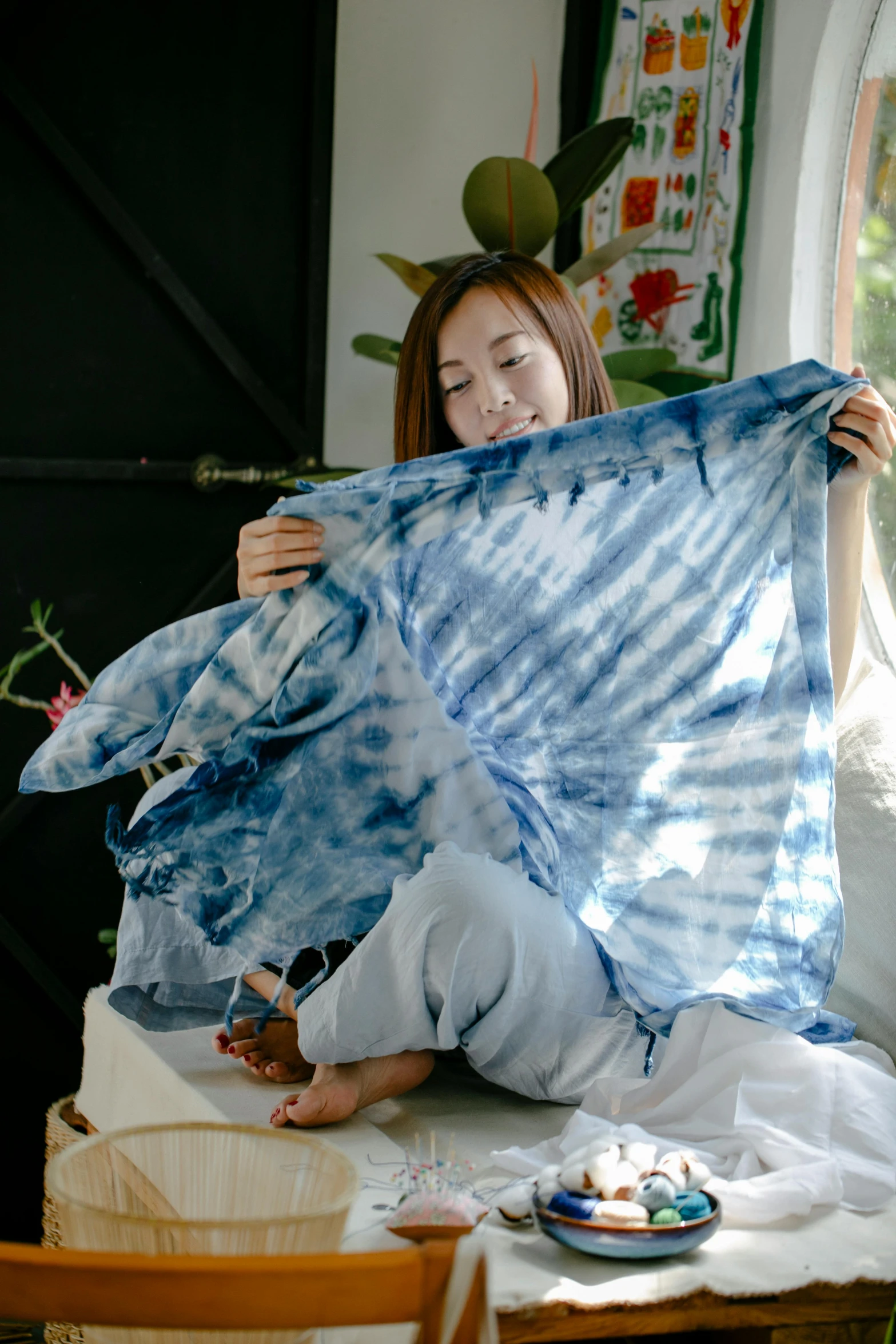 a woman sitting on a bed holding a blue tie dye shirt, inspired by Li Di, process art, folded, medium shot angle, ripped fabric, li zixin