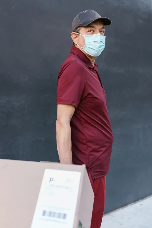 a man wearing a face mask and carrying a box, pexels contest winner, red shirt brown pants, in a dark teal polo shirt, profile image, (doctor)