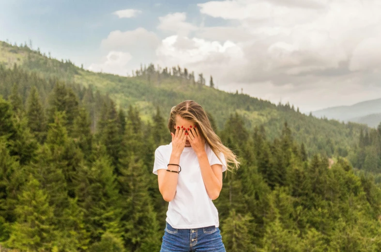 a woman standing on top of a lush green hillside, by Julia Pishtar, pexels contest winner, facepalm, jeans and t shirt, spruce trees, the girl is scared