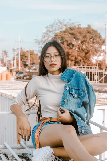 a woman sitting on a bench wearing a denim jacket, an album cover, inspired by helen huang, trending on pexels, wearing small round glasses, asian descent, looking confident, streetwear