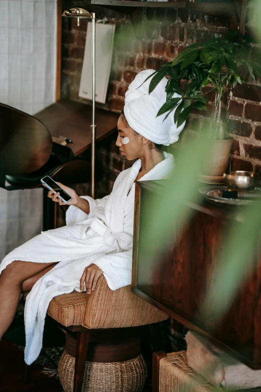 a woman in a bathrobe using a cell phone, by Everett Warner, trending on pexels, renaissance, hair styled in a bun, in a comfortable chair, spa, cloth head wrap