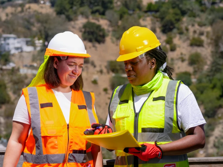 a couple of women standing next to each other, hivis, hill, holding a clipboard, mechabot