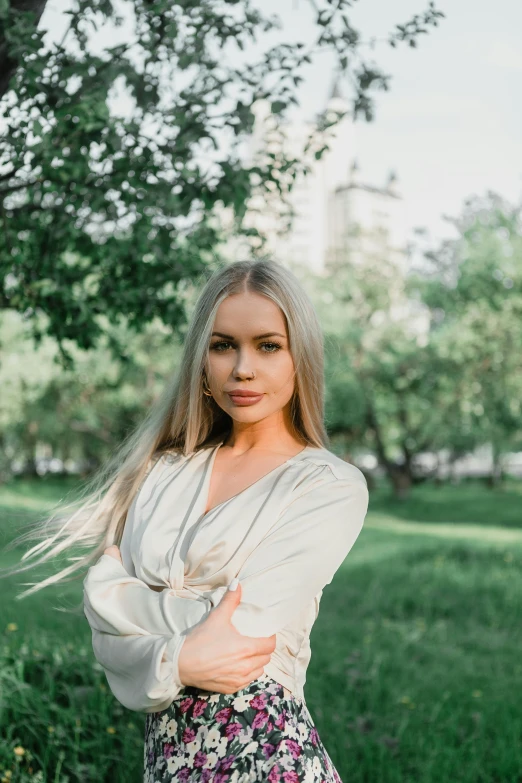 a woman standing in the grass with her arms crossed, pexels contest winner, blonde and attractive features, yulia nevskaya, in a city park, silver，ivory