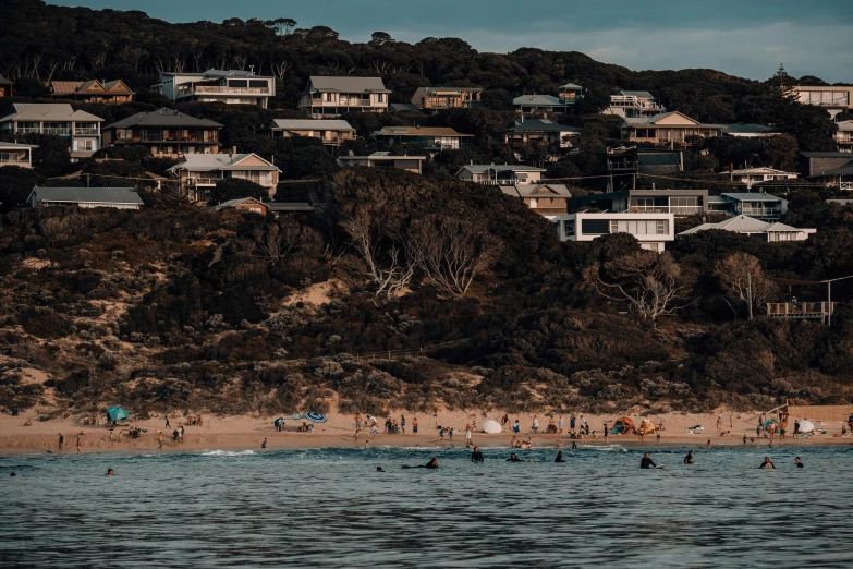 a group of people riding surfboards on top of a sandy beach, by Lee Loughridge, unsplash contest winner, waterfront houses, view from the sea, melbourne, the village on the cliff