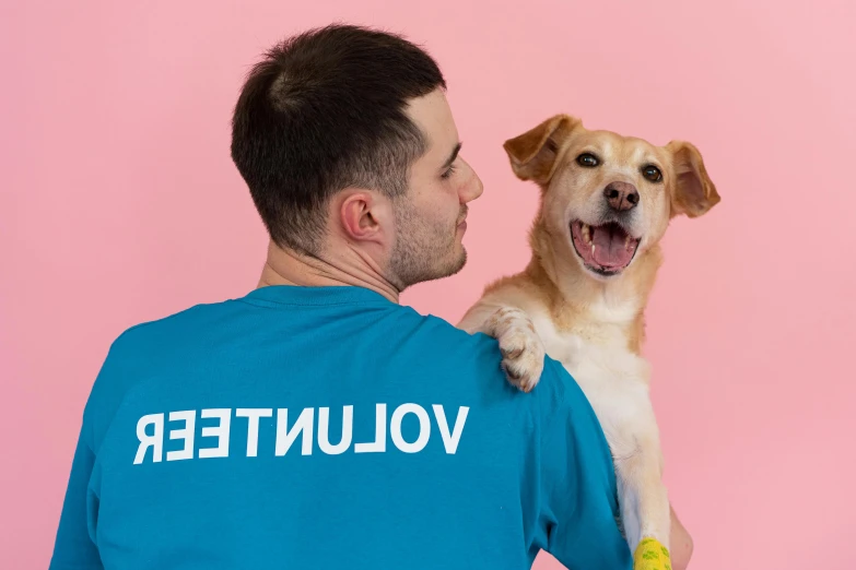 a man holding a dog wearing a volunteer t - shirt, a photo, aestheticism, pink, background image, hunched over, panting