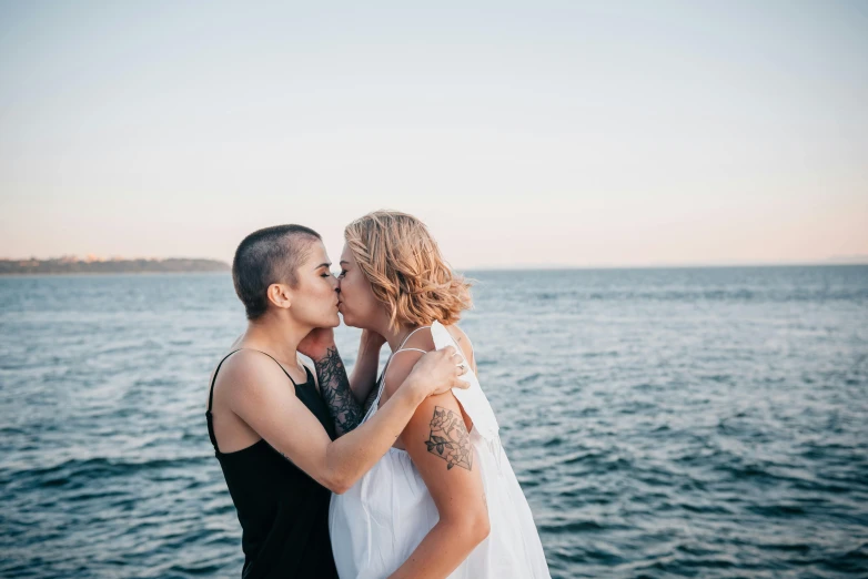 a man and a woman kissing in front of a body of water, a photo, by Liza Donnelly, unsplash, an epic non - binary model, viewed from the ocean, sydney hanson, two women
