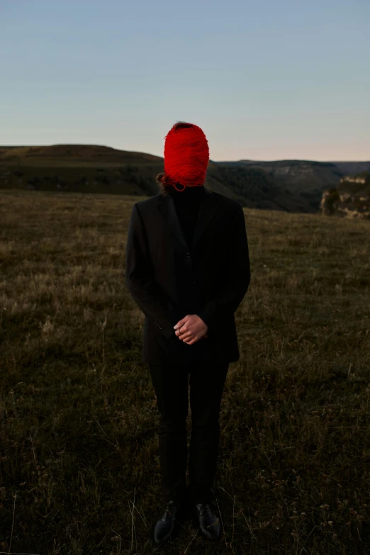 a person standing in a field with a red hat, an album cover, unsplash, balaclava mask, panoramic shot, lachlan bailey, late evening